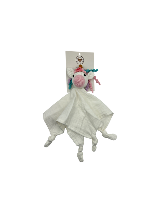 A white muslin comforter blanket featuring a crocheted unicorn head with a pink spiral nose, rainbow mane, and small pink and blue horns, attached to a white paper header card.